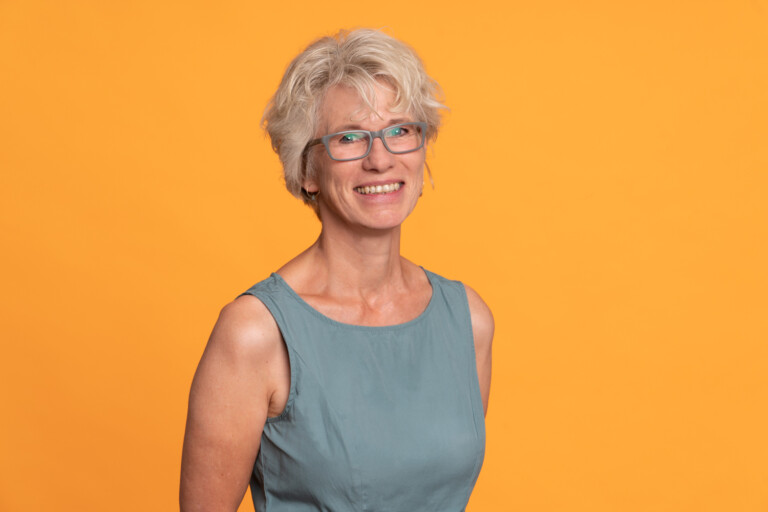 Porträt von Susanne Lenz in grün-grauem ärmellosen Oberteil vor orange-farbenem Hintergrund
