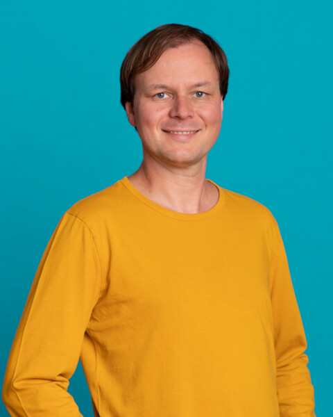 Porträt von Christoph Brammertz im gelben Pullover vor blauem Hintergrund