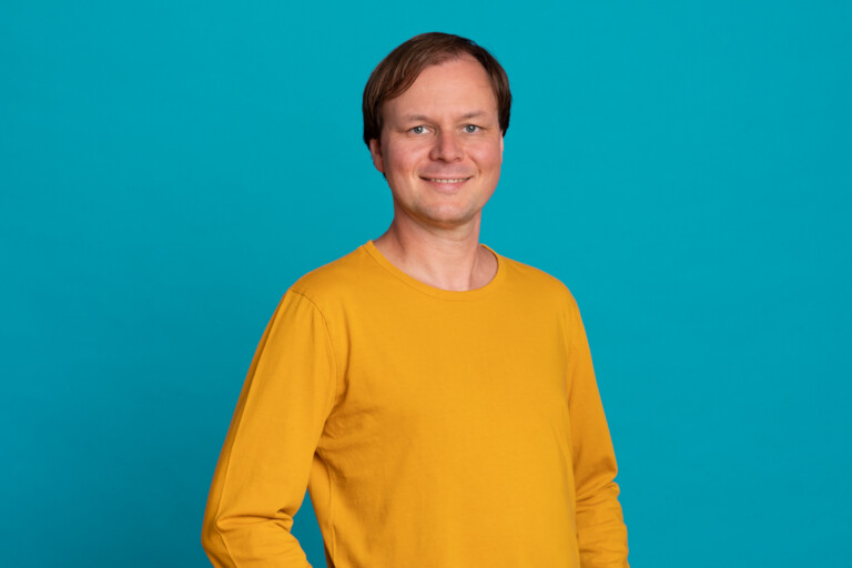 Porträt von Christoph Brammertz im gelben Pullover vor blauem Hintergrund