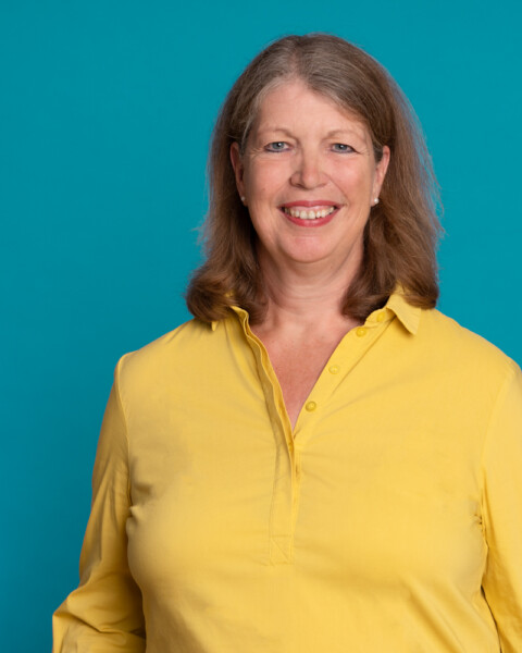 Porträtfoto von Almuth Fricke in einer gelben Bluse vor einem türkisfarbenen Hintergrund