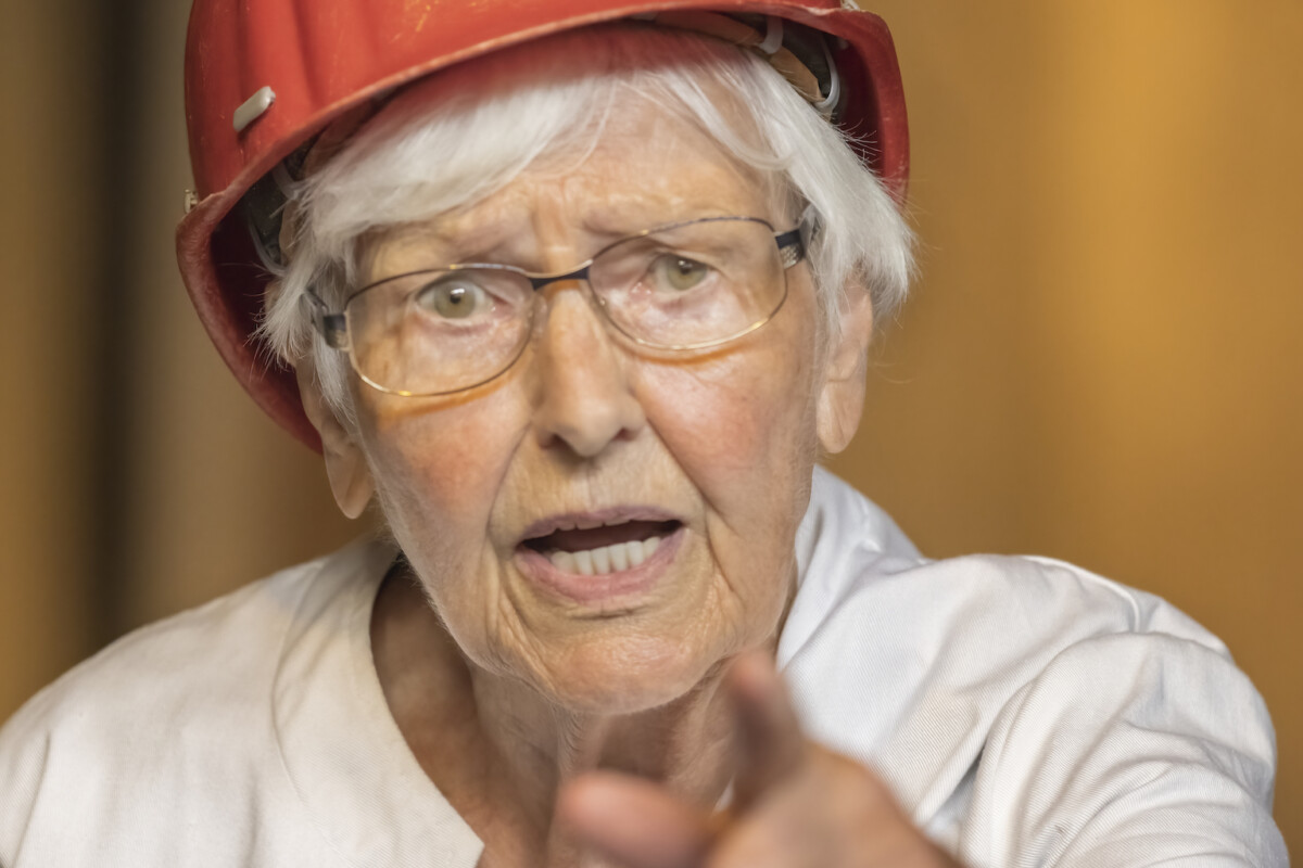 Eine ältere Frau mit rotem Schutzhelm zeigt mit dem Finger in die Luft. Sie hat Falten im Gesicht und trägt eine Brille. Ihr Mund ist leicht geöffnet, als wolle sie etwas Wichtiges sagen.