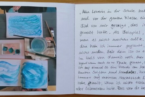 Ein aufgeschlagenes Heft zeigt auf der linken Seite ein Foto mit Farben, Pinseln, einem Becher und Aquarellen mit blauen wellenförmigen Motiven in Draufsicht. Auf der rechte Seite steht ein handgeschriebener Text.