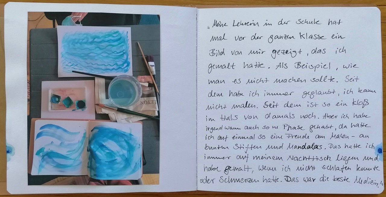 Ein aufgeschlagenes Heft zeigt auf der linken Seite ein Foto mit Farben, Pinseln, einem Becher und Aquarellen mit blauen wellenförmigen Motiven in Draufsicht. Auf der rechte Seite steht ein handgeschriebener Text.