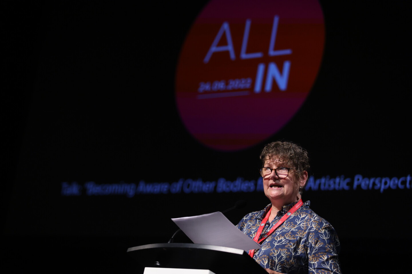 Keynote-Sprecherin Kaite O´Reilly am Rednerpult unter einem All-In-Schriftzug