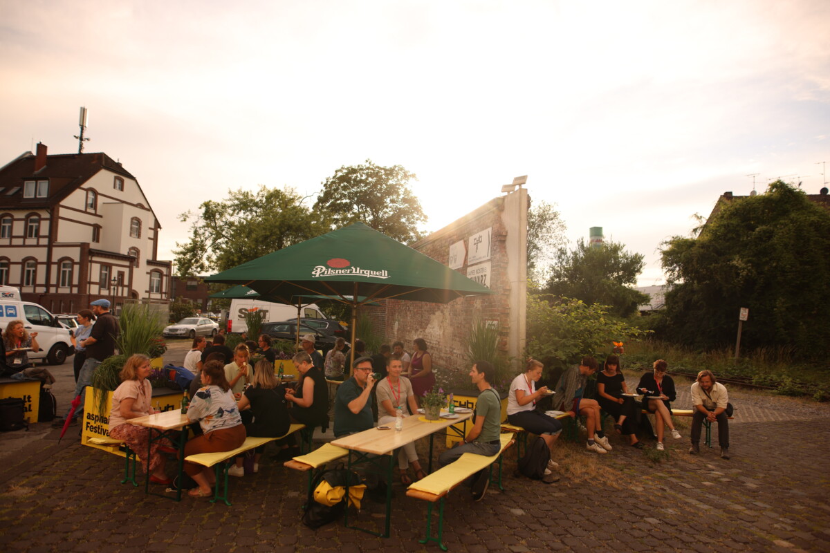 Totale eines alten Fabrikgeländes mit mehreren Biertischen und -bänken mit einem großen Sonnenschirm darüber, an denen einige Menschen sitzen.
