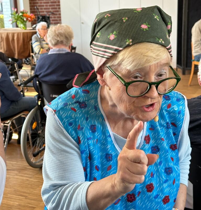 Eine ältere Frau in blauer Kittelschürze mit Brille, Kopftuch und erhobenem Zeigefinger
