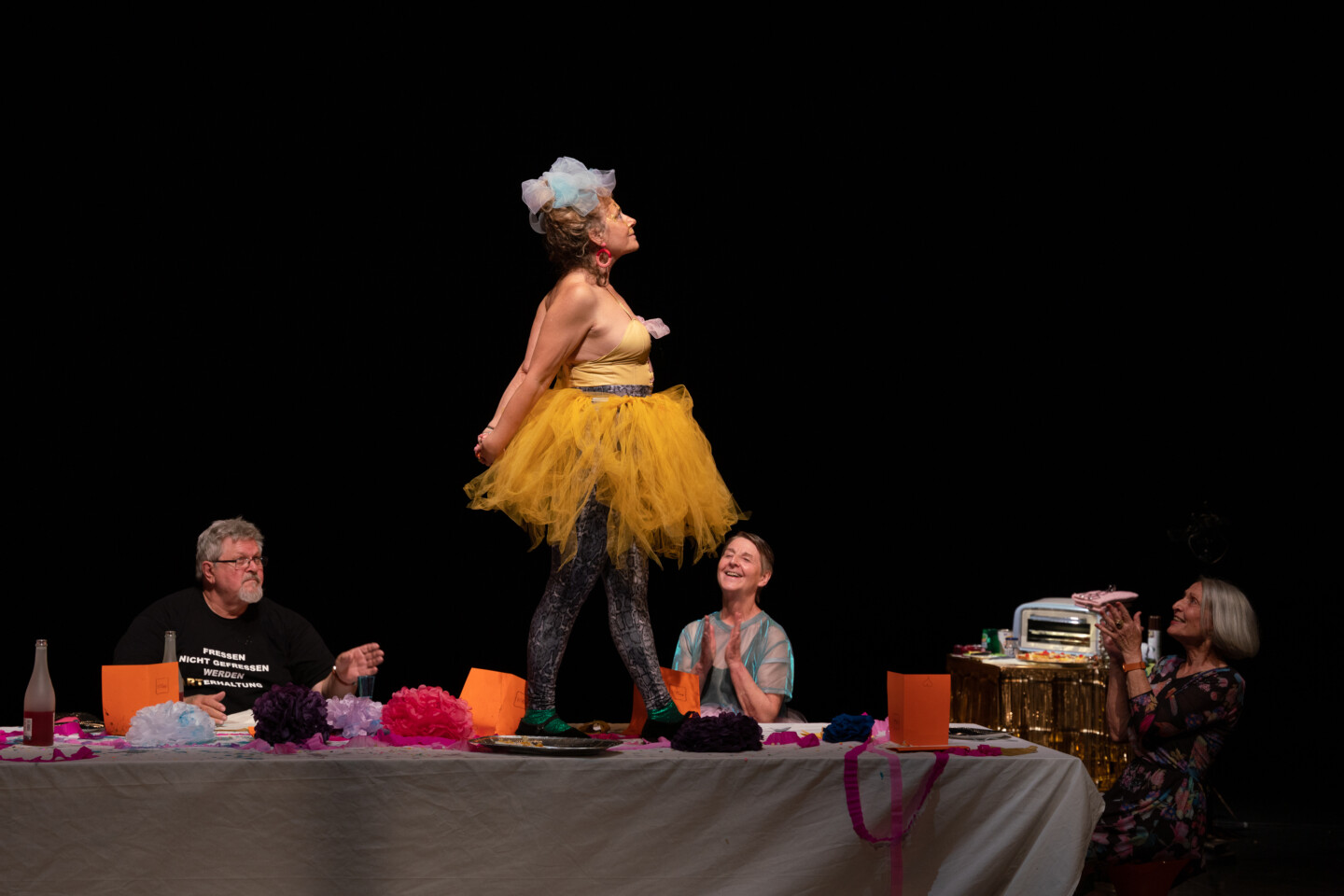 Bühnenfoto aus "Wir hatten die Zeit unseres Lebens" von Stefan Mießeler. Eine grauhaarige Frau in einem gelben Kleid mit Tütü, die auf einem Tisch steht, um den herum Menschen sitzen und ihr applaudieren.