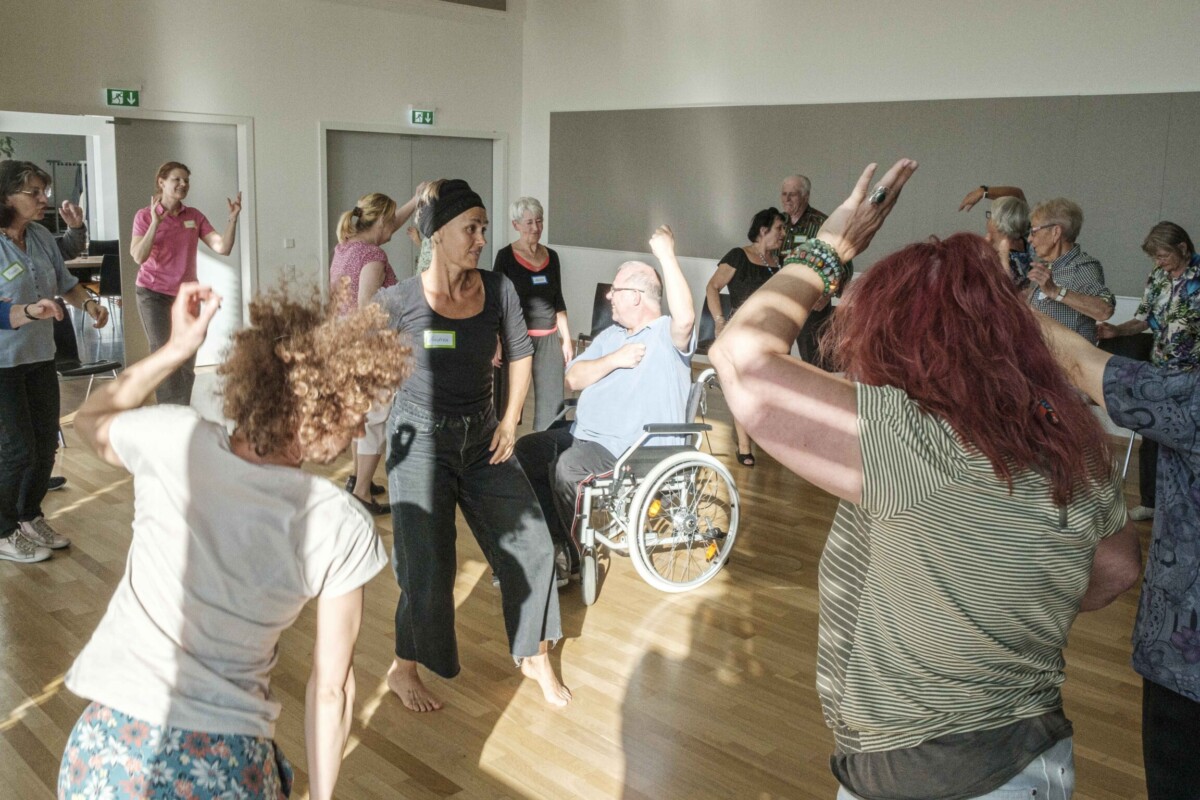 A group of people of different ages dance through a room flooded with sunlight. In the centre of the picture is a man in a wheelchair who has also raised his arms in a dancing movement.