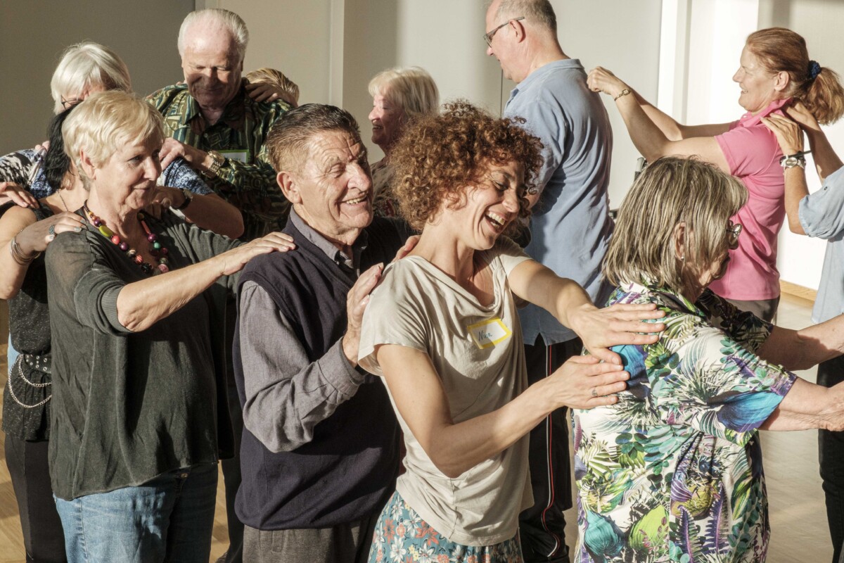 Eine Gruppe fröhlicher, meist älterer Menschen tanzt in einer Art Polonaise durch den Raum.