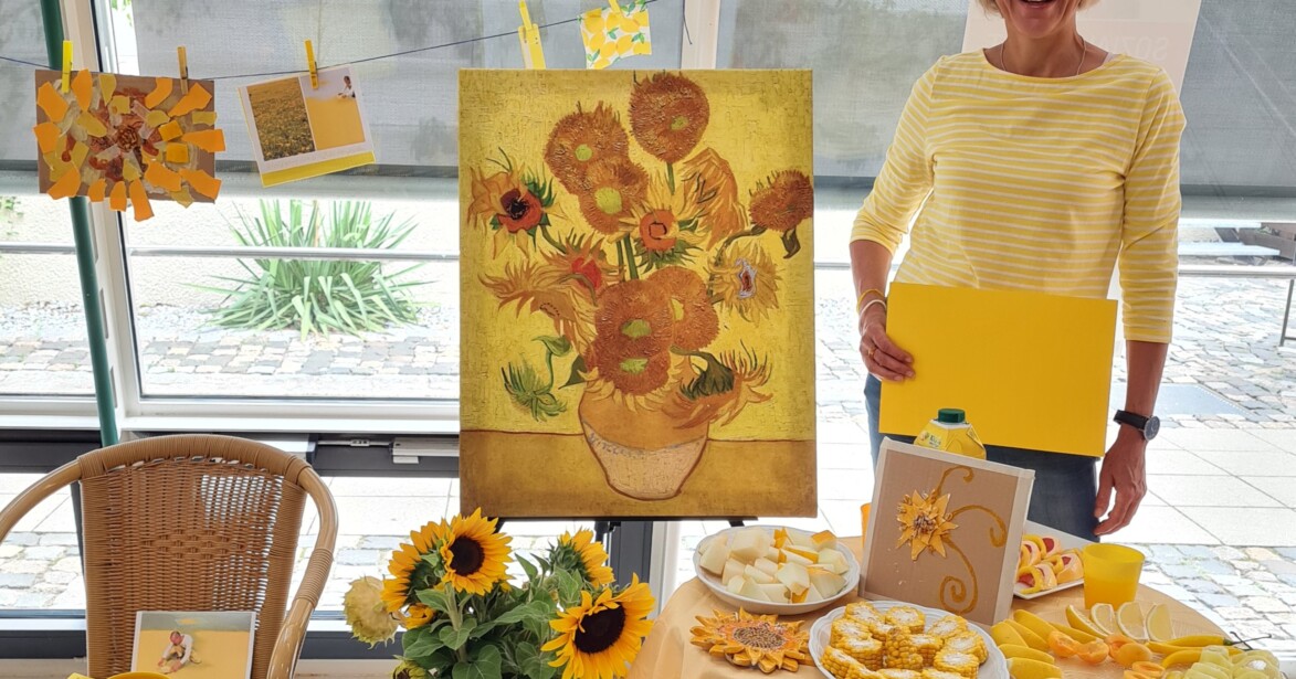 Petra Kellermann steht zwischen Gegenständen zur Farbe Gelb und einem Gemälde mit Sonnenblumen