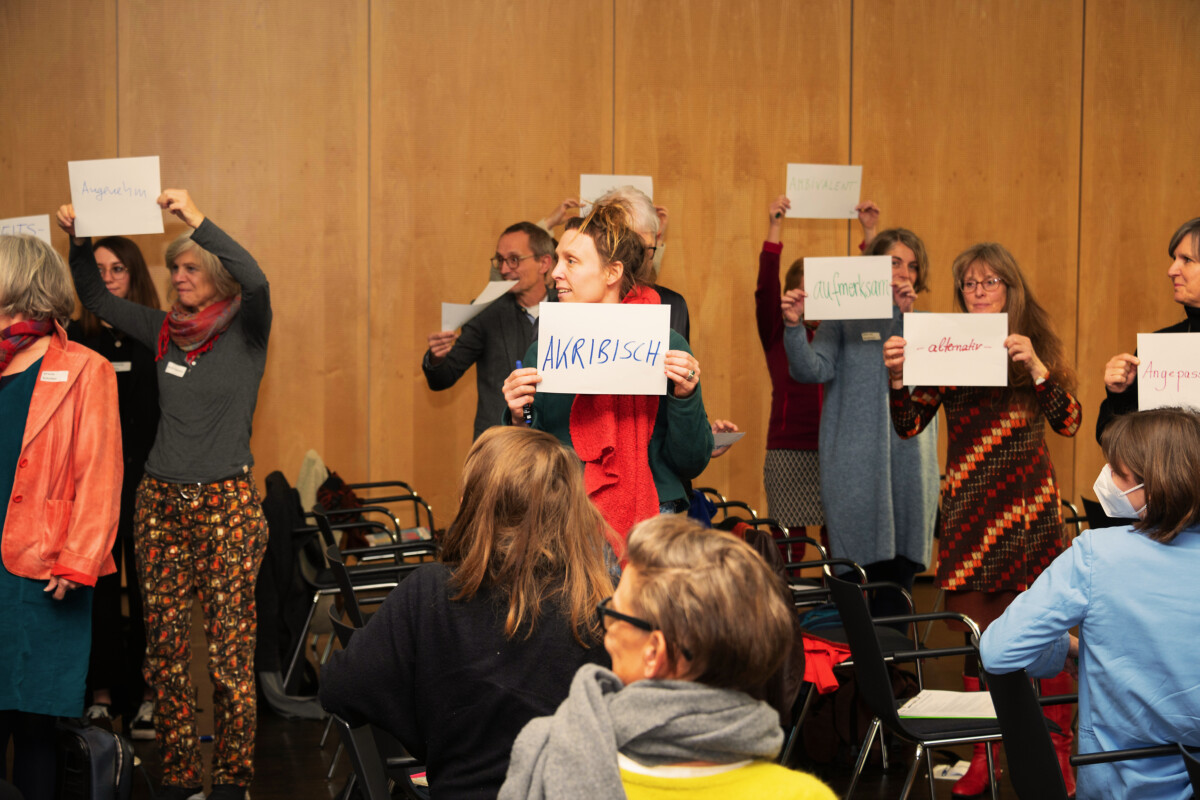 Participants in the cultural geragogy course stand in an event hall holding up signs. In the center is a woman with the word "akribisch" (meticulous) handwritten on her sign.