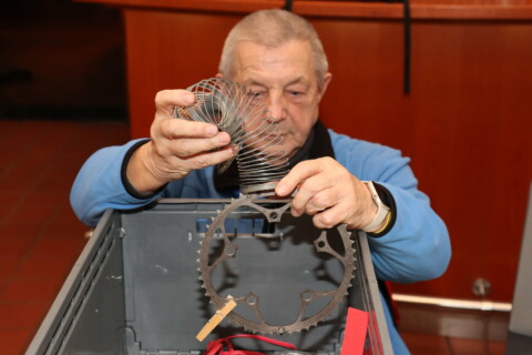Ein älterer Mann im blauen Arbeitskittel hält eine Metallfeder in der Hand.