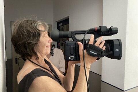 Eine ältere Frau hält eine große Videokamera in den Händen und guckt in den Sucher.
