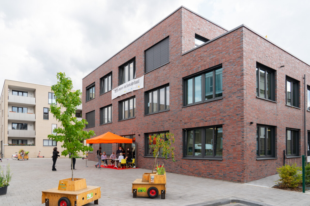 A modern, three-storey brick building with a flat roof. In front of it are mobile wooden seating areas, plants and a parasol. The photo was taken at the opening of the Seekabelhaus 2021.