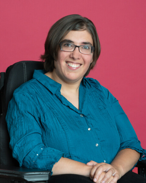 Portrait of Isabell Rosenberg, smiling, with dark hair and glasses in wheelchair, against magenta background