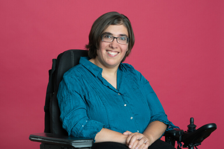 Porträt von Isabell Rosenberg, lächelnd, mit dunklem Haar und Brille im Rollstuhl. vor magentafarbenem Hintergrund