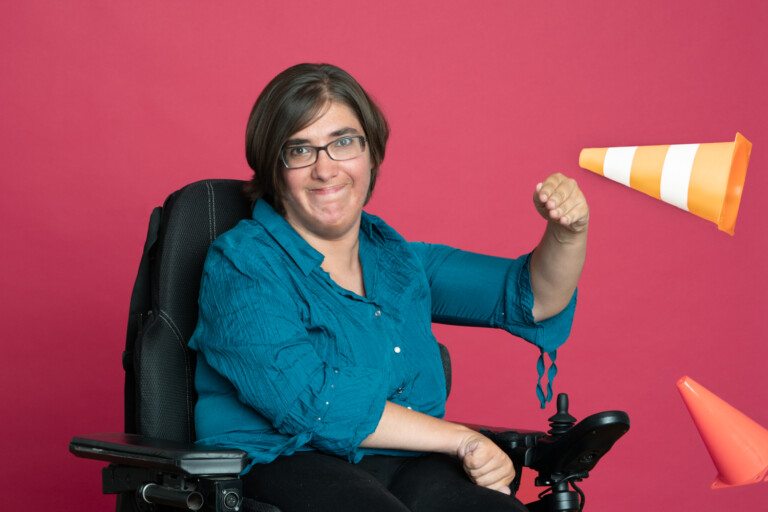 Porträt Isabell Rosenberg mit dunklem Haar und Brille im Rollstuhl, die rot-weiße Verkehrspylone wegschlägt