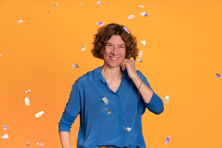 Woman with curly hair wearing blue shirt in front of orange background. It is raining banknotes.