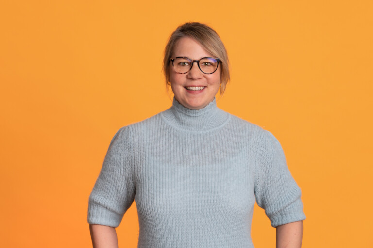 Portrait of Annette Ziegert with glasses, dark blond hair and a grey turtleneck jumper in front of an orange background