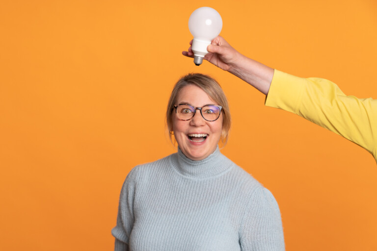 Portrait of Annette Ziegert against an orange background. She is wearing glasses and a green-grey turtleneck jumper. Above her head a hand holding a light bulb