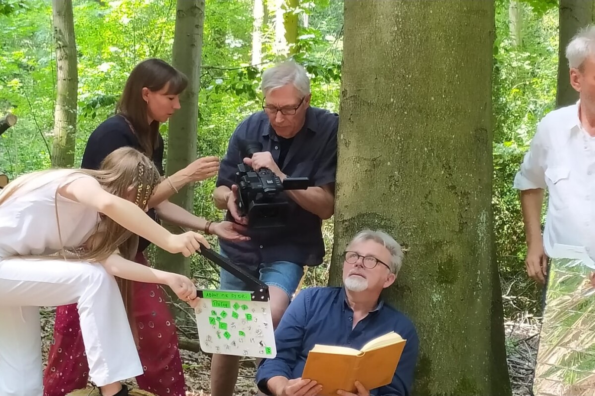 Ein älterer Mann sitzt mit einem Buch in der Hand an einen Baum gelehnt auf dem Boden. Ein anderer älterer Mann filmt ihn dabei von schräg oben. Neben dem Kameramann steht eine Frau, die ihm andere Kamera etwas zeigt. Im Vordergrund steht eine Jugendliche, die eine Filmklappe bedient.