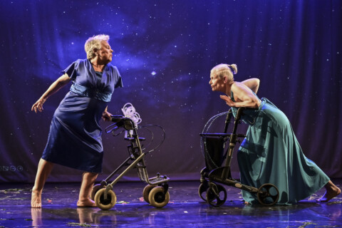 Two elderly women stand on a stage in dresses, each leaning on a walker.