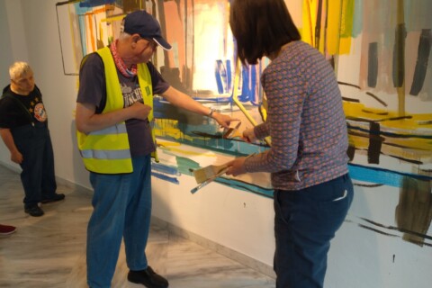Ein älterer Mann mit gelber Warnweste zeigt mit seiner linken Hand auf ein Gemälde an der Wand. Eine Frau rechts von ihm einen Pinsel. Im Hintergrund steht ein zweiter älterer Mann