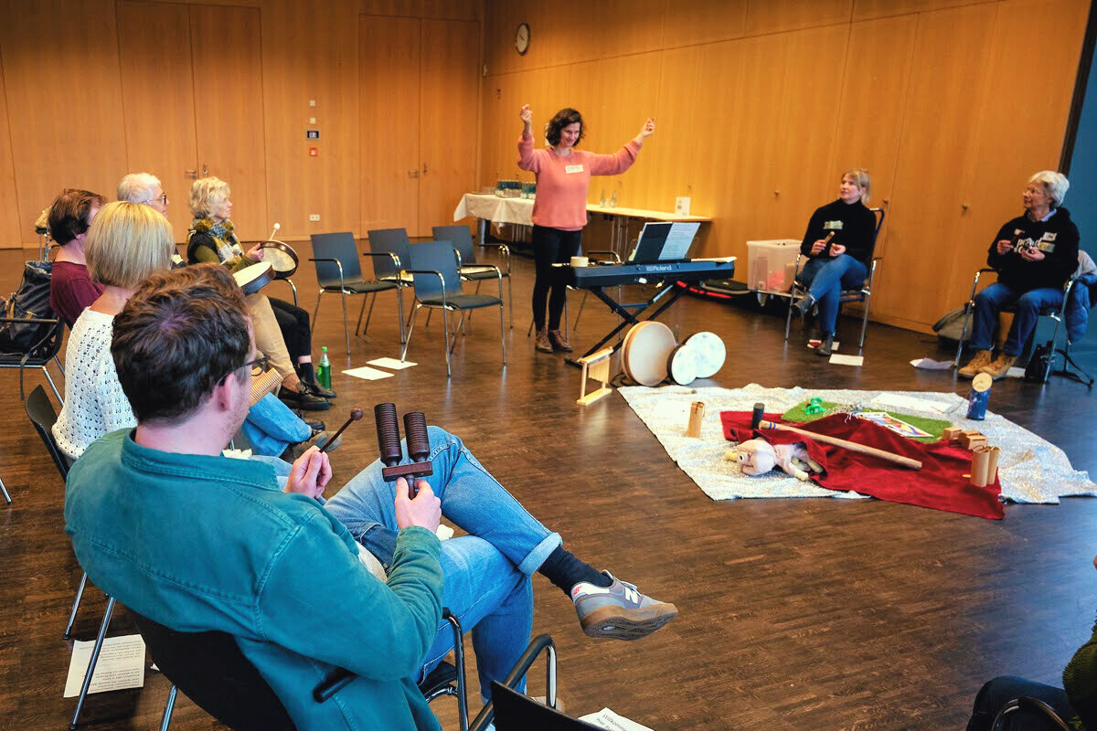Eine Gruppe von Personen, die auf Stühlen im Kreis sitzten und auf eine Fau blicken, die mit erhobenen Armen ähnlich einer Dirigentin vor ihnen steht. Auf dem Boden in der Mitte liegen verschiedene Musikinstrumente und Requisiten.