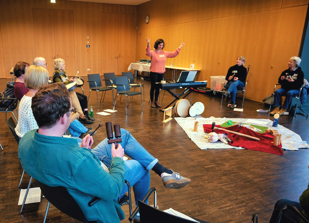 Eine Gruppe von Personen, die auf Stühlen im Kreis sitzten und auf eine Fau blicken, die mit erhobenen Armen ähnlich einer Dirigentin vor ihnen steht. Auf dem Boden in der Mitte liegen verschiedene Musikinstrumente und Requisiten.