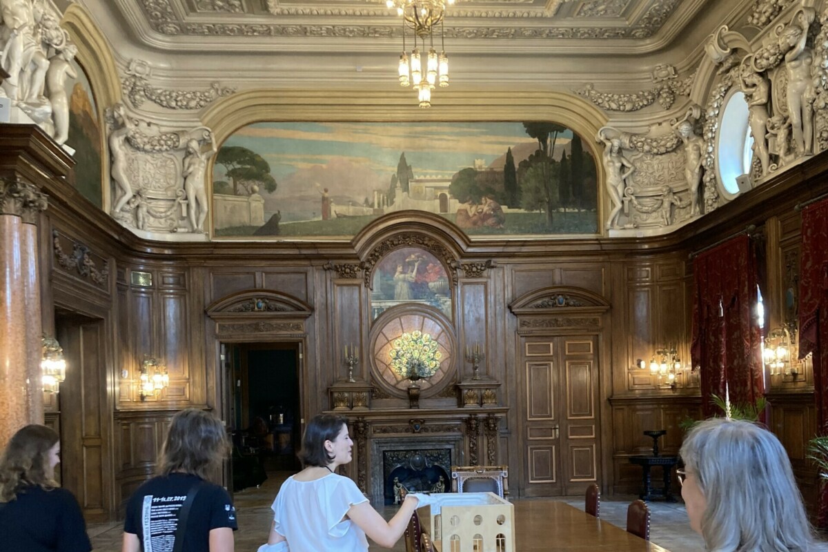 A group of people in a high room richly decorated with wood panelling and stucco with a large table in the middle on which a model of the room is placed.