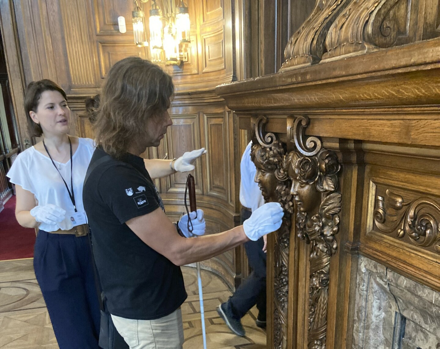 A woman wearing white gloves stands next to a man holding a cane for the blind and also wearing white gloves. He is touching a brass-coloured relief in the shape of a head on a richly decorated mantelpiece.