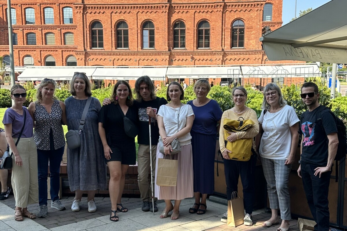 A group of people posing for a photo in front of an old red brick building. One person has a blind man's stick in his hand.