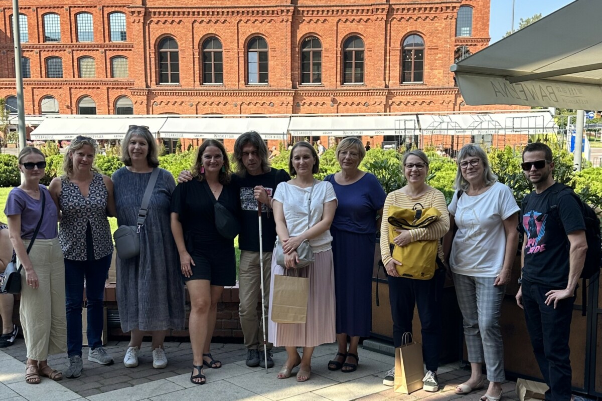 Eine Gruppe von Menschen posiert für ein Foto vor einem alten roten Backsteingebäude. Eine Person hält einen Blindenstock in der Hand.