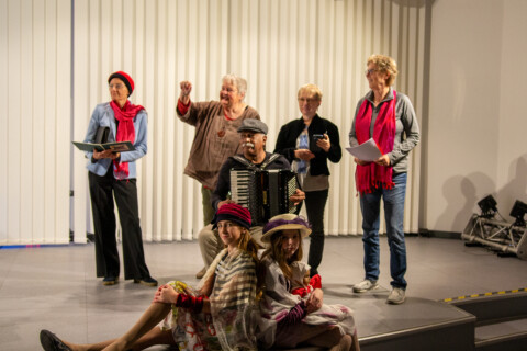 Zwei Mädchen mit Hüten und einer Stola bekleidet, sitzen an einem Bühnenrand. Hinter ihnen sitzt ein älterer Mann mit einem Akkordeon. Dahinter stehen vier ältere Frauen. Die äußeren halten Hefte in ihren Händen.