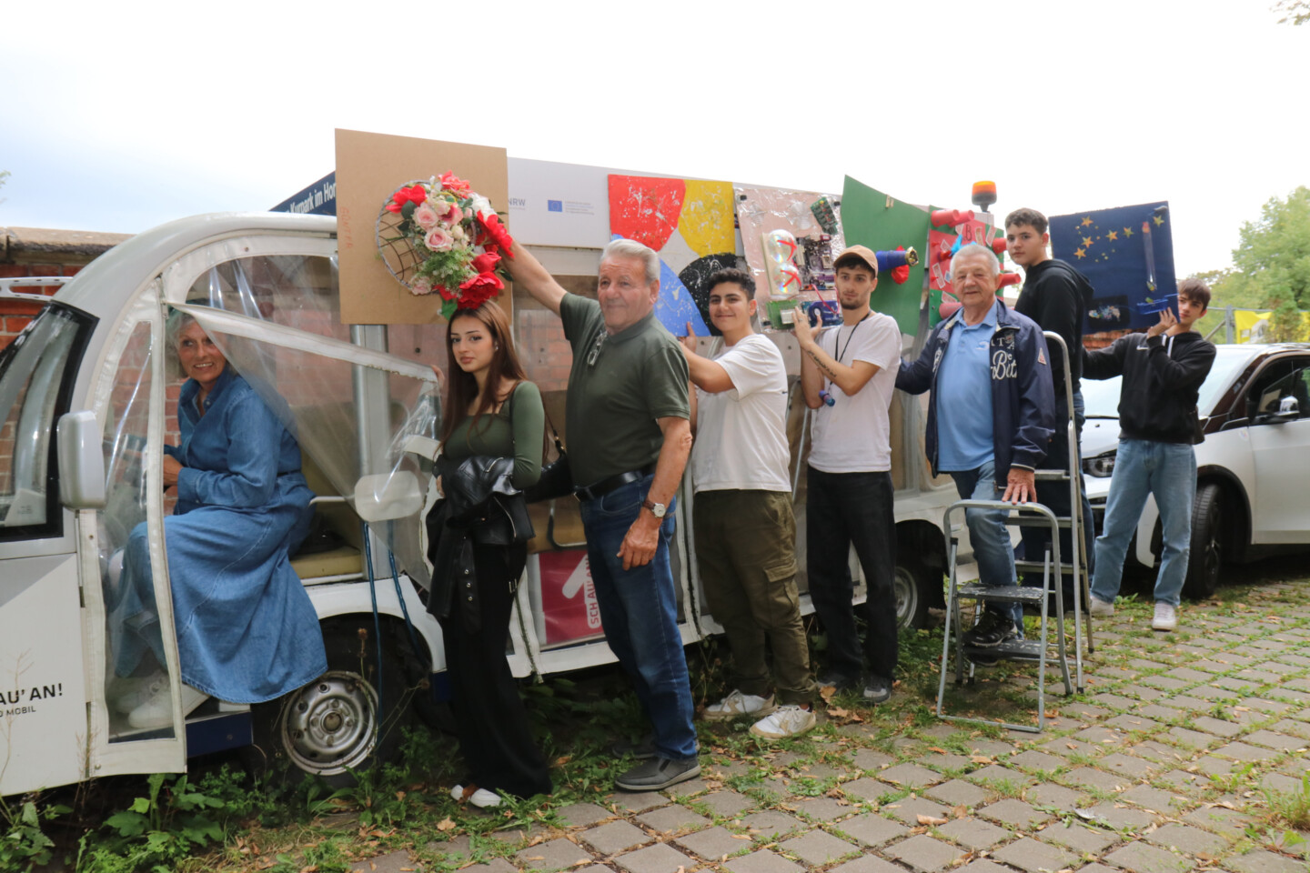 Jugendliche und Ältere stehen vor einem Stretch-TukTuk, das mit Blumen und verschiedenen gebastelten Kunstwerken geschmückt ist