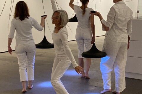 Vier weißgekleidete Frauen bewegen sich durch einen weißen Raum. Drei tragen jeweils eine Lampe, die blaue Punkte auf den Boden wirft.