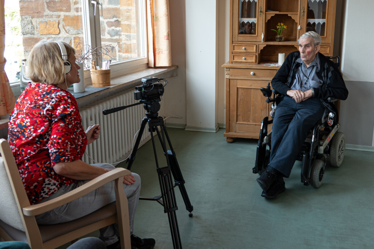 Eine Frau mit Kopfhörern filmt einen Mann, der auf einem Rollstuhl sitzt.
