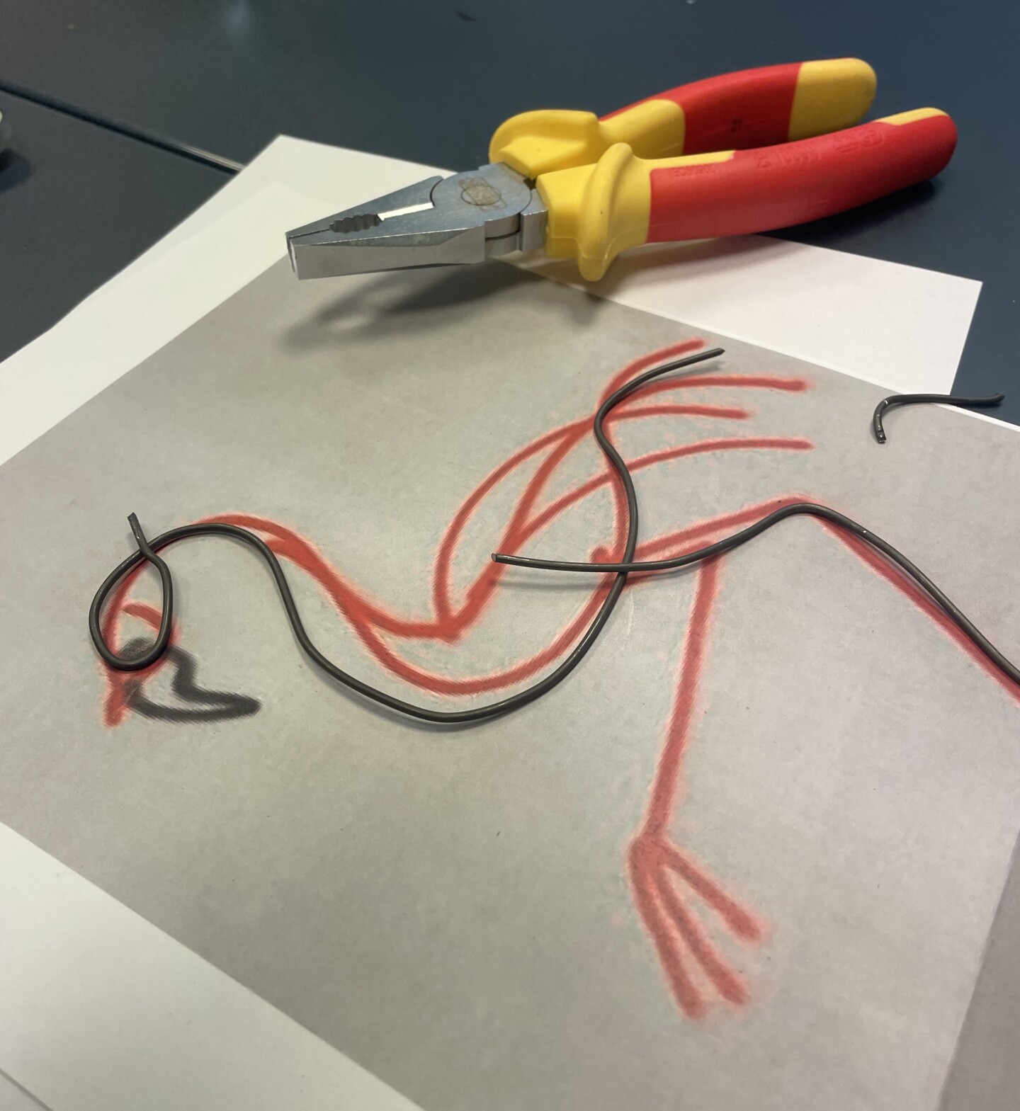 Eine Zange und Draht, der zur Form eines Vogel gebogen wurde, auf einem Blatt Papier, auf dm die Vogelform vorgezeichnet wurde.