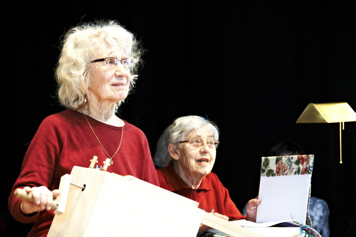Zwei ältere Frauen Frauen in roten Oberteilen. Die eine hat einen Holzkasten mit einer Kutbel vor sich.