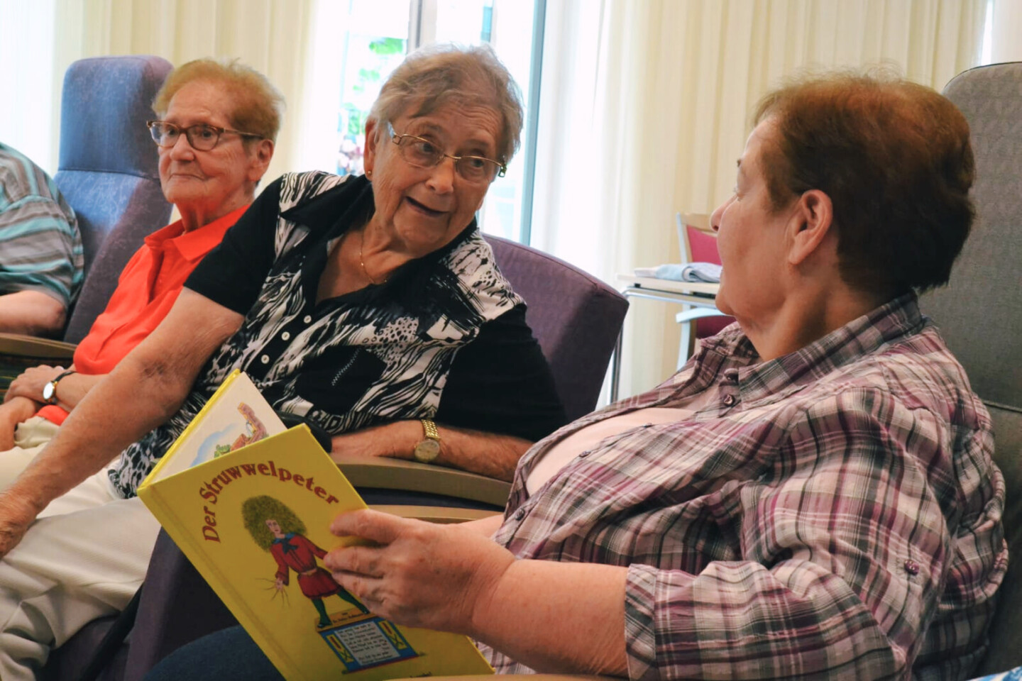 Drei ältere Frauen sitzen nebeneinander. Die Frau im Vordergrund hält ein aufgeschlagenes Struwwelpeter-Buch in den Händen.