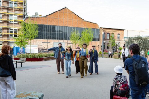 Fünf Personen verschiedenen Alters in Kleidung im Stil der 1970er Jahre laufen in Richtung der Kamera über eine Freifläche vor alten Fabrikgebäuden und machen unterschiedliche Bewegungen mit ihrem rechten Arm.
