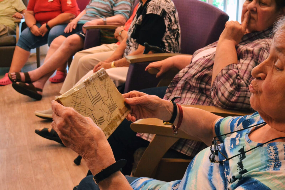 Eine Gruppe von älteren Menschen, die auf Stühlen sitzen. ein Frau im Vordergrund schaut auf eine alte Pappschachtel, auf der in verschnörkelter Schrift „Schach, Dame, Mühle“ steht.