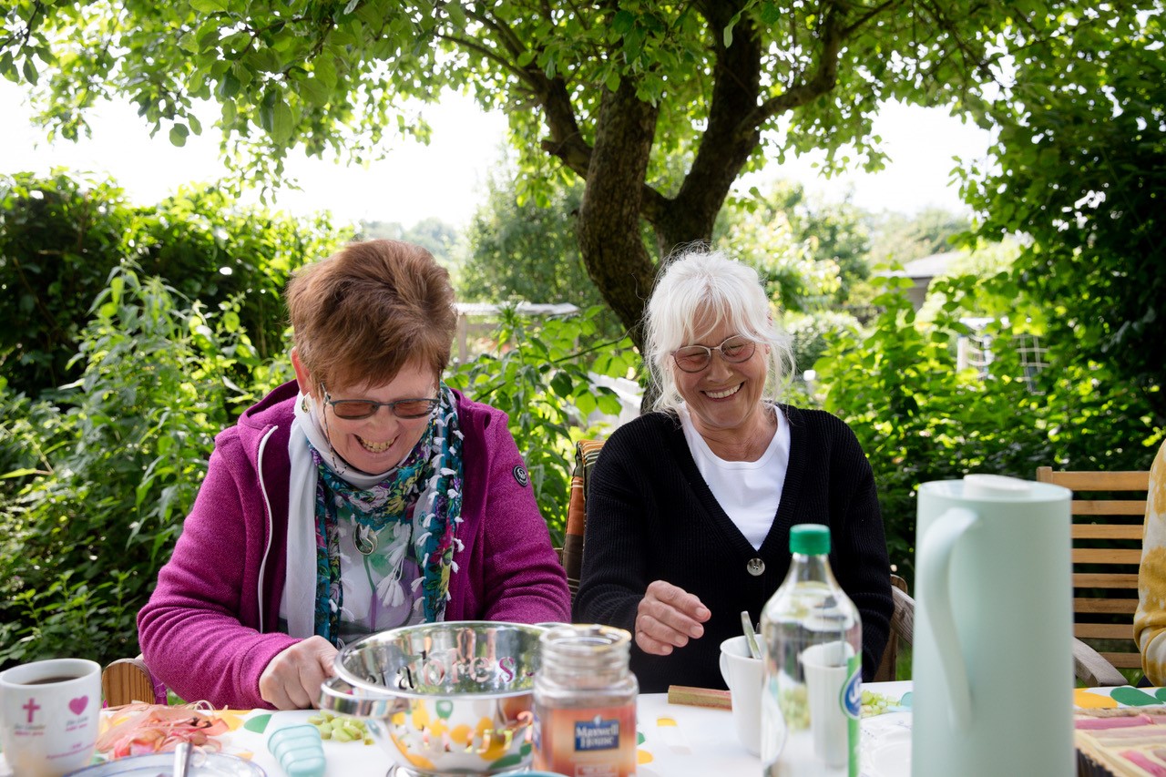 Zwei ältere Frauen sitzen lachend an einem Tisch unter einem Baum im Garten. und schneiden Gemüse.