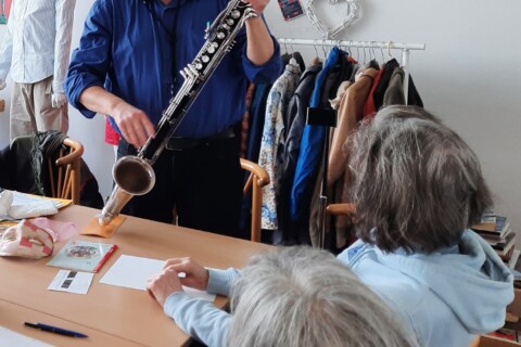 Ein Mann steht mit einer Klarinette vor zwei Frauen, die an einem Tisch sitzen.