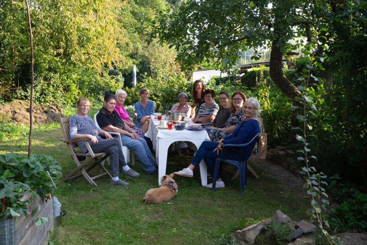 Eine Gruppe von zehn Personen sitzt an einem weiß gedeckten Tisch in einem grünen Garten. Sie sind entspannt, lächeln und unterhalten sich, während Getränke und Speisen auf dem Tisch stehen. Ein kleiner Hund liegt auf dem Rasen in der Nähe.
