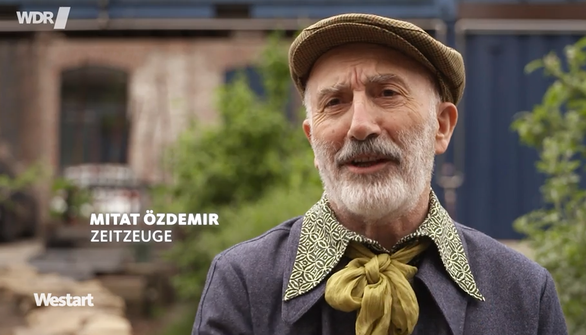 Screenshot aus einem TV-Beitrag, der einen älteren Mann mit Bart, Schiebermütze und ein Halstuch zeigt. Im Bild steht "Mitat Özdemir, Zeitzeuge / WDR Westart"