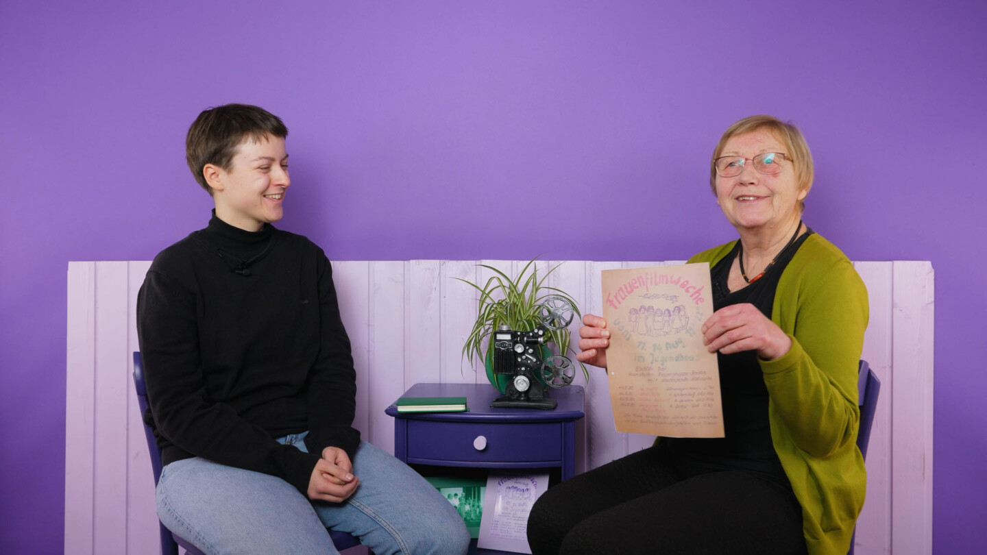 Vor einem lila Hintergrund sitzen Jutta Bringmann (r.), Mitbegründerin des Frauenfilmclubs Borken und eine junge Frau, die sie interviewt hat. Bringmann zeigt das handgemalte Plakat der ersten Frauenfilmwoche.