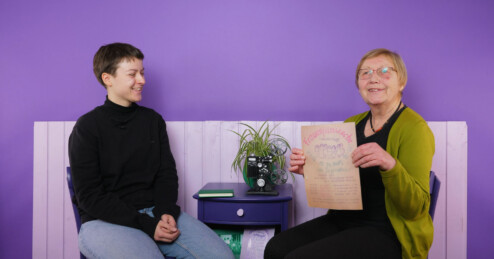 Vor einem lila Hintergrund sitzen Jutta Bringmann (r.), Mitbegründerin des Frauenfilmclubs Borken und eine junge Frau, die sie interviewt hat. Bringmann zeigt das handgemalte Plakat der ersten Frauenfilmwoche.
