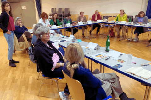 Gruppe von Personen sitzt und steht um einen U-förmig gestellten Tisch. Auf dem Tisch liegen bzw. stehen Papier, Stifte, Wasserfaschen und Namensschilder.