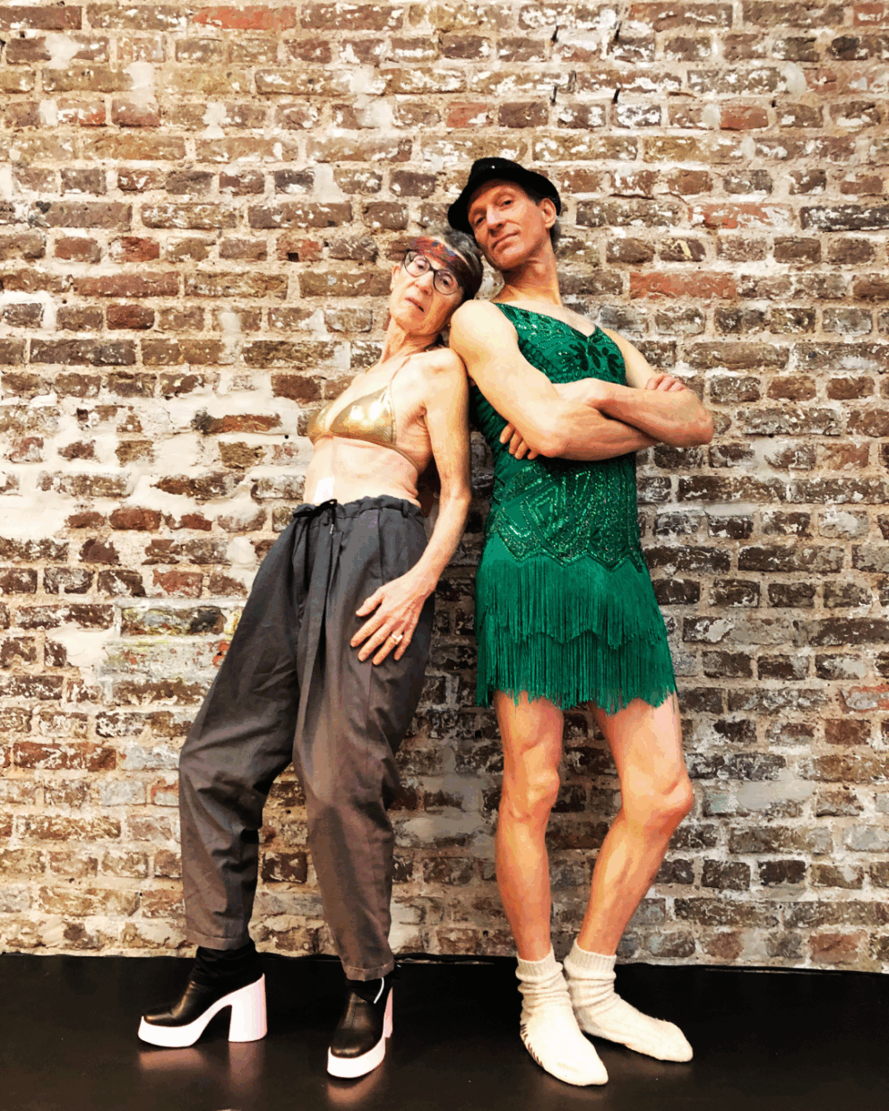 Zwei ältere Personen stehen vor einer Backsteinwand. Die linke Person trägt ein goldenes Bikini-Oberteil, graue Hosen und schwarze Plateauschuhe. Die rechte Person trägt ein grünes Kleid mit Fransen und weiße Socken.